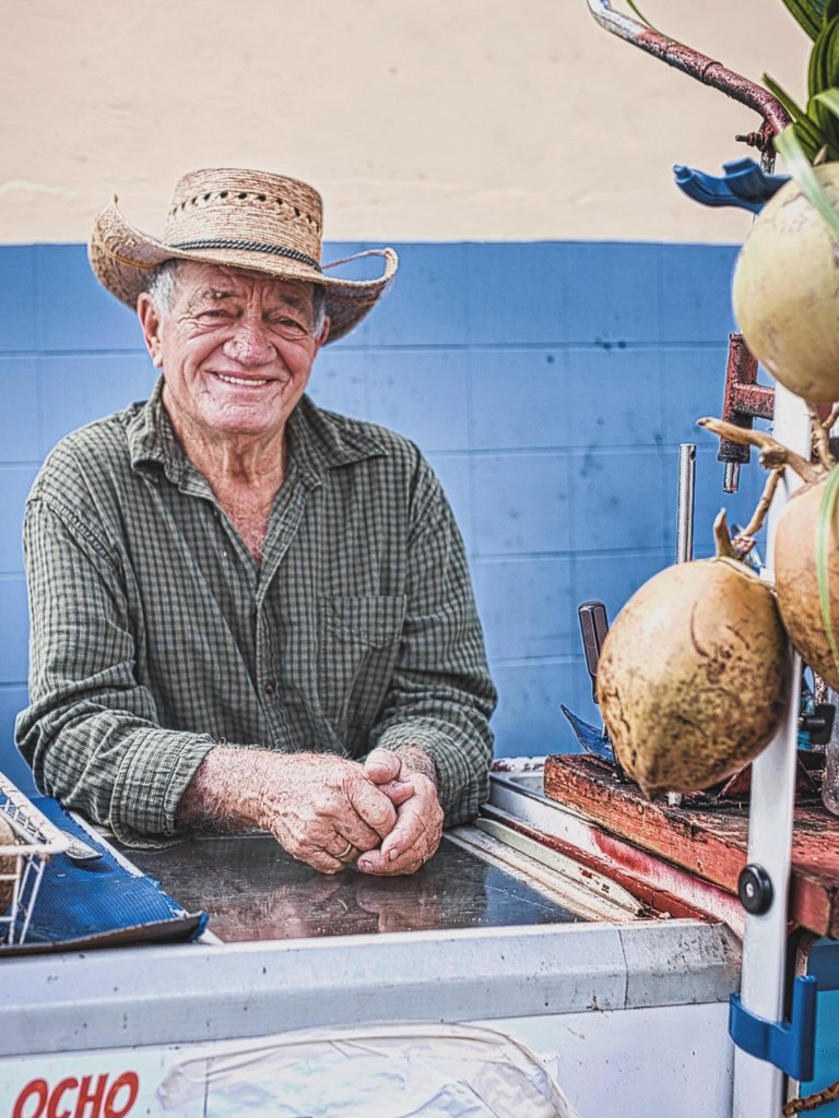 A gentleman behind a small coconut stand in Nassau, straw hat on, hands folded on the counter, watching the street.